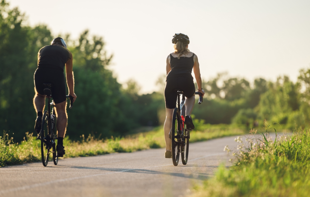 Two people riding bicycles on a road in the countryside surrounded by parks and trails.