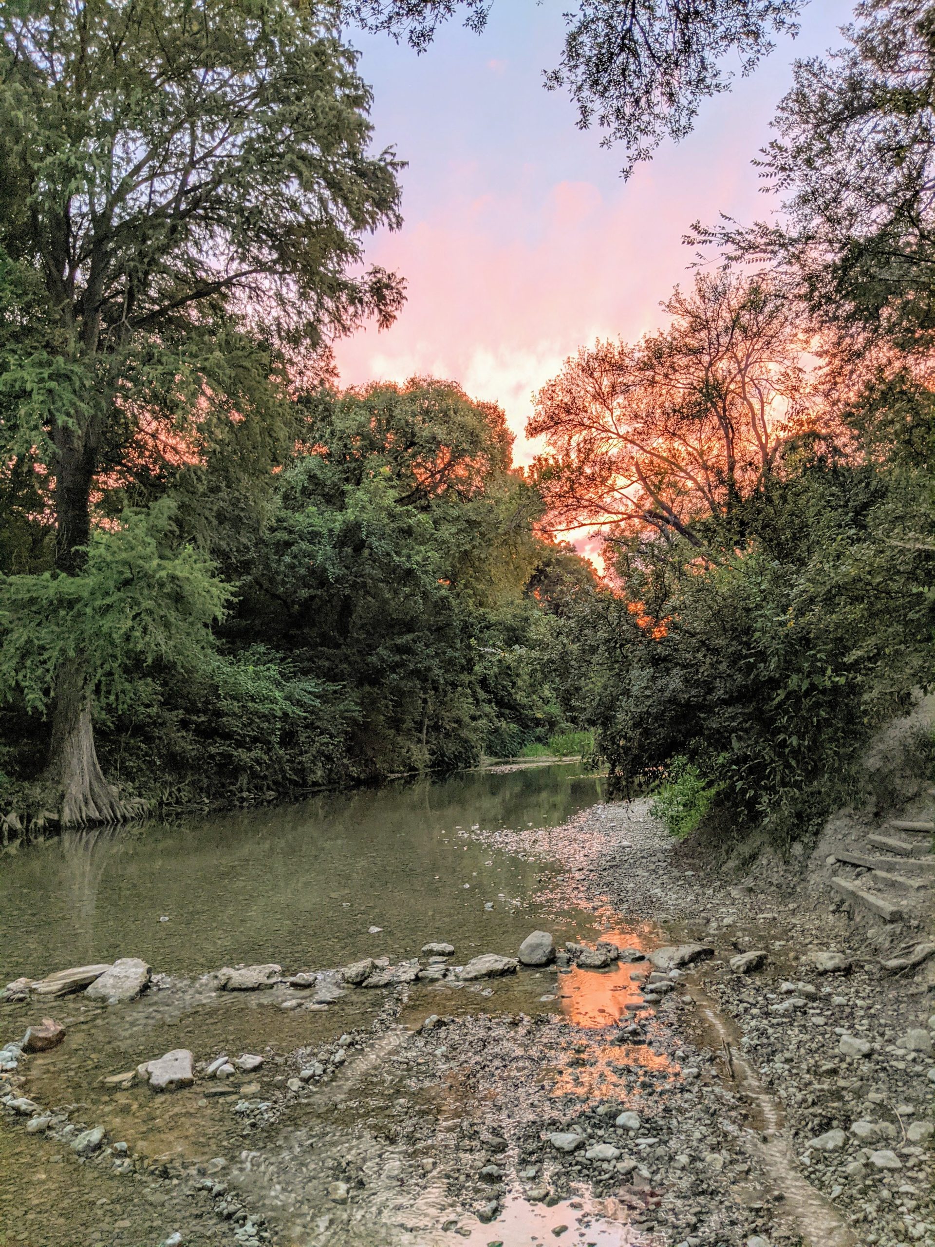New homes in New Braunfels Texas nestled alongside a scenic river flowing through lush parks and trails, adorned with rocks and trees, illuminated by the striking hues of a captivating sunset.