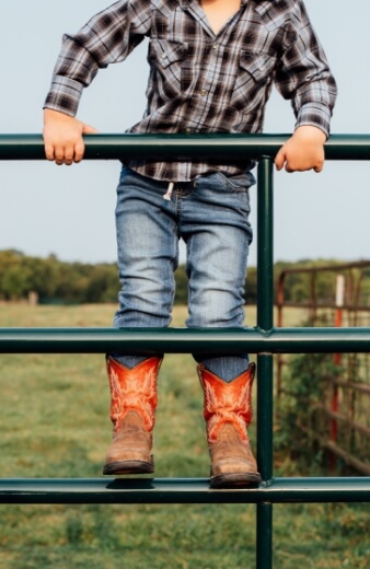 A little boy in cowboy boots leaning on a fence near parks and trails in Mayfair homes, New Braunfels Texas.