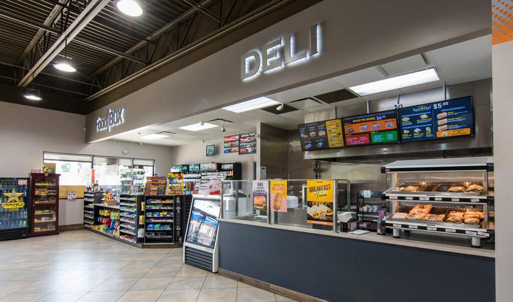 A convenience store deli counter displays hot food items, with menu boards above and shelves of snacks, drinks, and groceries in the background.