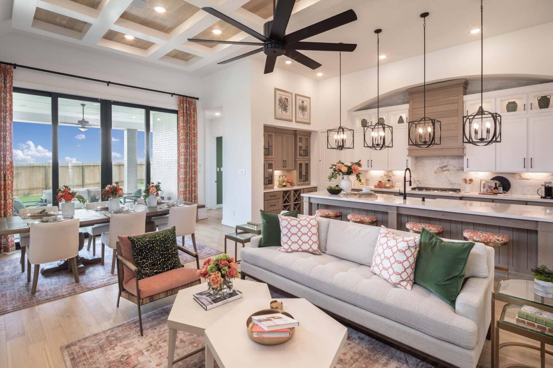 Open-concept living room and kitchen with modern furnishings, pendant lights, a ceiling fan, and large windows overlooking a fenced backyard.
