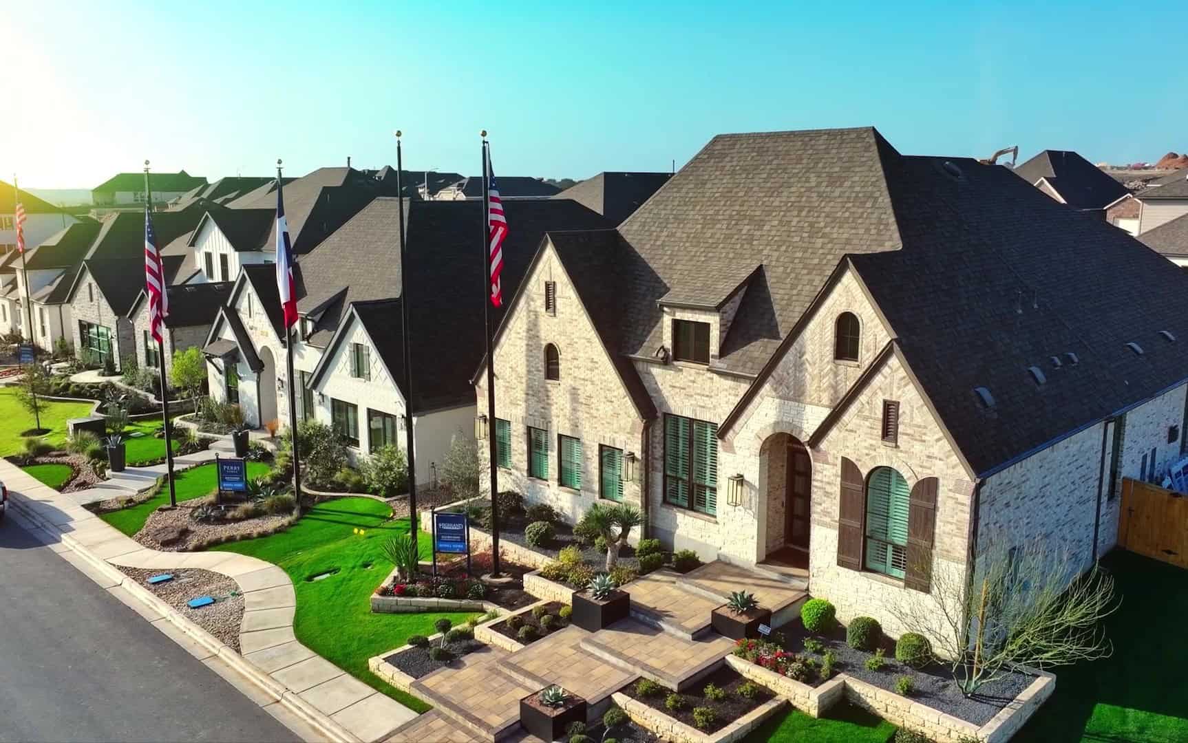 Row of modern homes in Mayfair New Braunfels community with manicured lawns, flags displayed in front, under a clear blue sky.