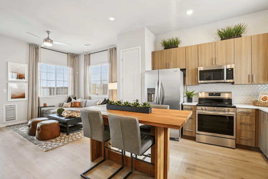 Modern open-concept kitchen and living room with wood cabinetry, stainless steel appliances, dining table, neutral decor, large windows, and potted plants.