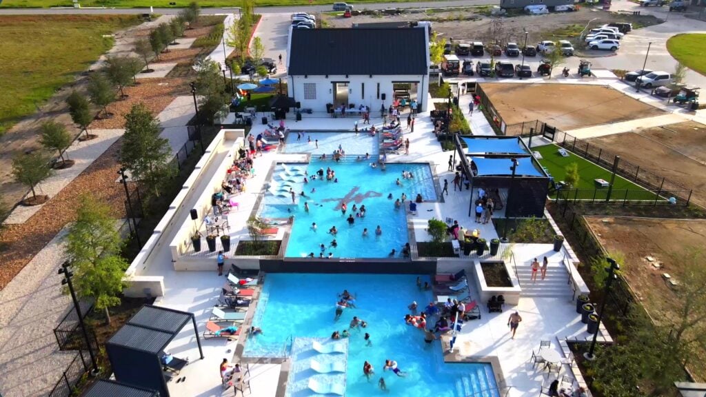 Aerial view of a busy outdoor pool area with people swimming and lounging, surrounded by chairs and umbrellas, with a building and parked cars in the background.