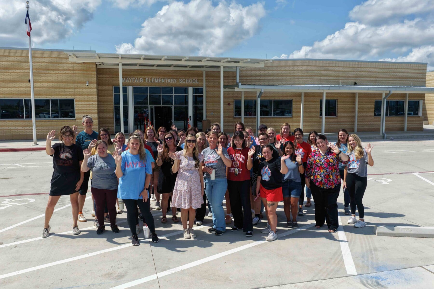 A group of teachers stands and waves outside Mayfair Elementary School on a sunny day, with the school building and a flagpole visible in the background.