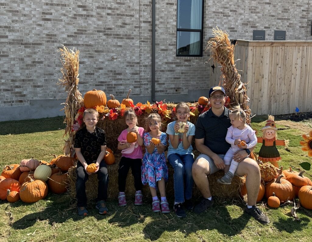 A family smiles while sitting on a hay bale. Orange pumpkins are seen all around the photo op area. Best Pumpkin Patches and Fall Events Near Mayfair